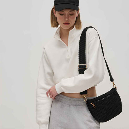 A model carrying a black woven crossbody vegan bag  with a wide black strap and gold-tone hardware against a light gray studio background.