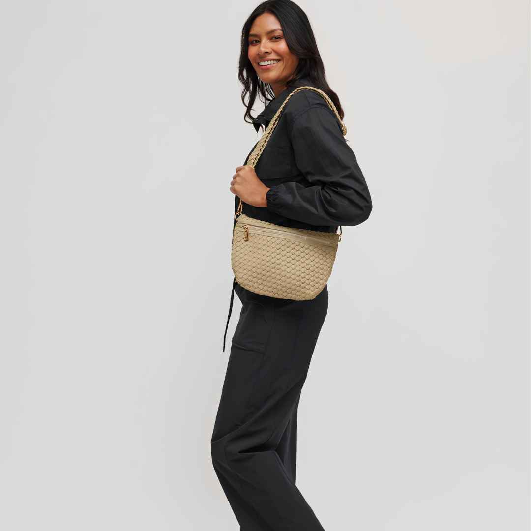 A woman carrying a woven beige vegan shoulder bag with a gold chain strap against a light gray studio background.