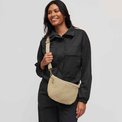 A woman wearing  a beige woven vegan crossbody bag with a matching strap against a light gray studio background.