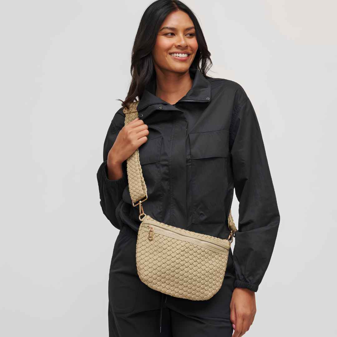A woman wearing a beige woven crossbody vegan bag with a matching woven strap against a light gray studio background.